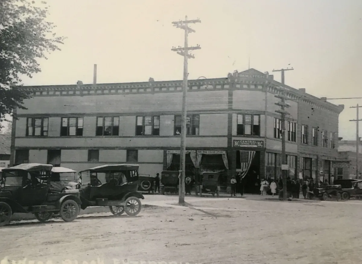 A lively street scene in downtown Enterprise features a beautifully preserved historic building as the centerpiece. Vintage cars are parked along the curb, and groups of people are seen congregating on the sidewalk, suggesting a vibrant, active community. The architecture evokes a sense of history and connection to the town's past, while the bustling atmosphere captures the dynamic spirit of the area.