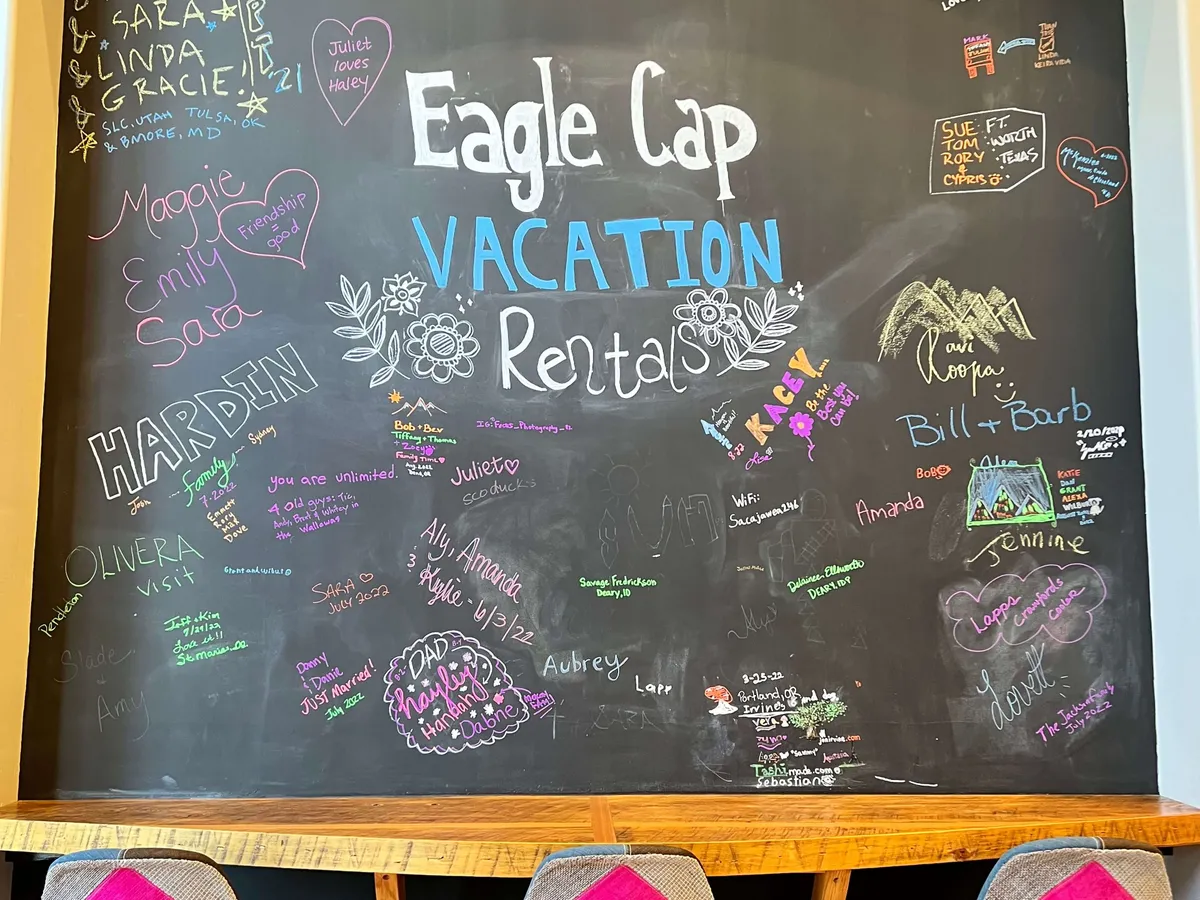 A colorful, welcoming chalkboard in a communal interior space, filled with guest messages, vibrant drawings, and the text 'Eagle Cap VACATION Rentals.' The space invites reading, sharing, and a sense of belonging.