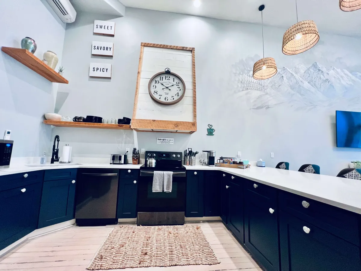 A cozy kitchen and dining area adorned with a large clock, wooden shelves displaying decorative items, handy countertop appliances, and warm lighting. A rug underfoot adds texture and warmth, and the setting radiates comfort, making it ideal for gathering, cooking, and sharing meals with friends or family.