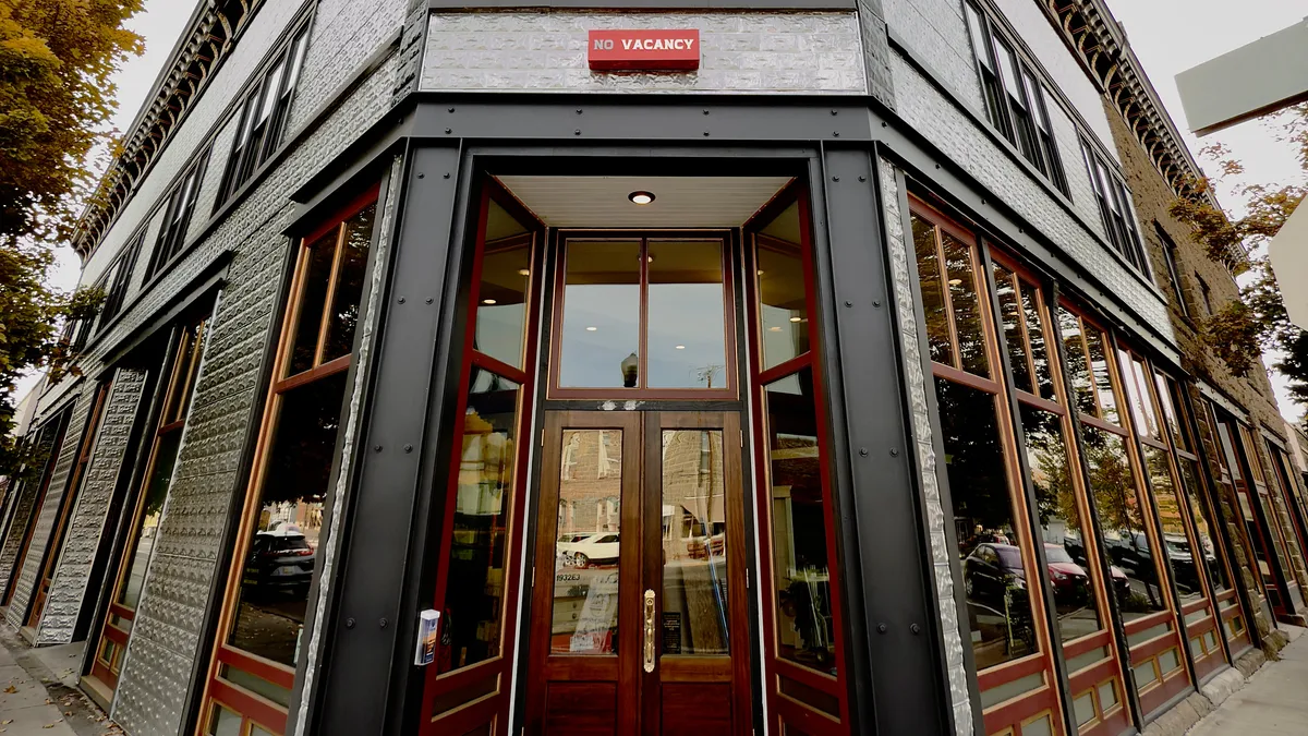 This image captures the main entrance of a building with large windows, a visible 'NO VACANCY' sign, and scenes of people visiting nearby shops, dining, and socializing. Outdoor seating and signs of local commerce highlight the vibrant activity right outside the entrance.