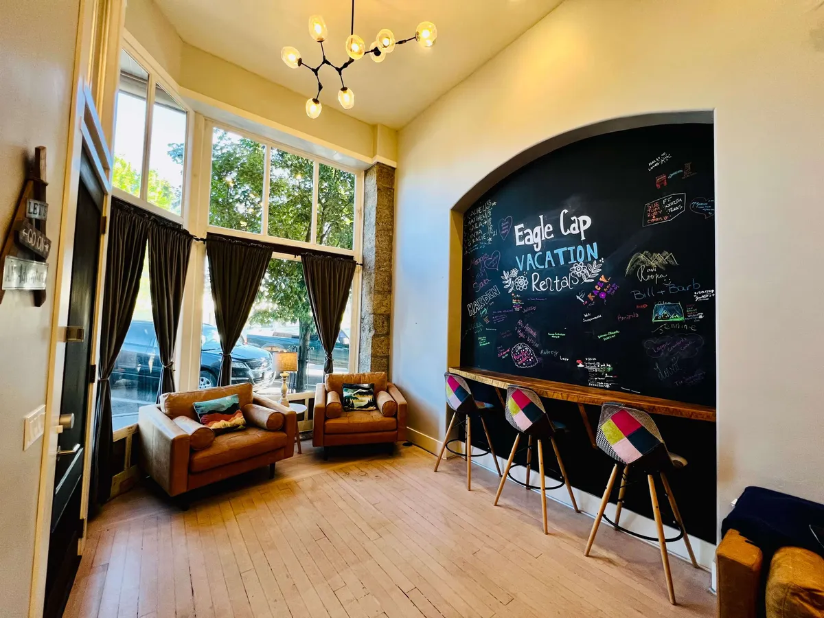 A communal area featuring a chalkboard wall filled with colorful messages, two inviting armchairs, a bar seating area with vibrant stools, and large windows draped with curtains. The space is designed for relaxed socializing, casual drinks, and fostering a sense of belonging.