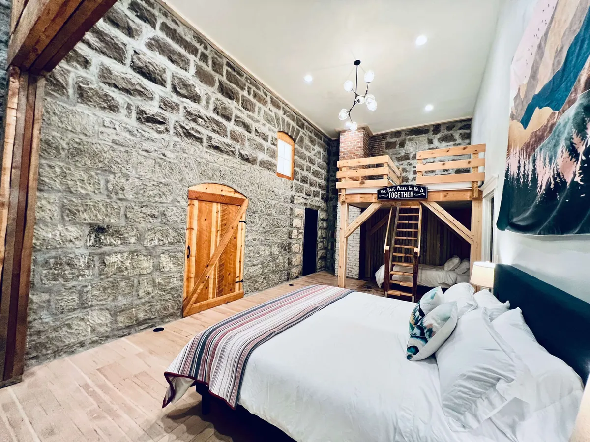 The image showcases the condo's inviting interior, featuring a lofted bed area with plush bedding, rustic stone walls, a charming wooden door, and tasteful decorative accents. Warm lighting creates a cozy atmosphere, suggesting a perfect space for relaxing, sharing stories, or gathering with family. The blend of historic details and modern comfort is immediately apparent, making the space both unique and welcoming.