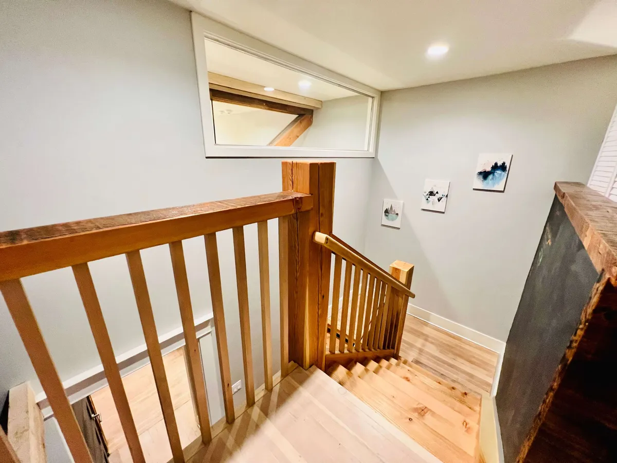 This image features a warm, inviting interior hallway with a wooden staircase and framed art adorning the walls. A large window lets in natural light, drawing the eye to both the art and the inviting atmosphere of the space.