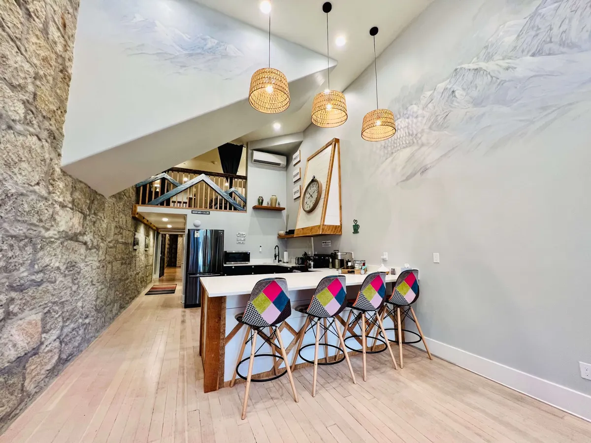 A bright, contemporary kitchen featuring a central island with colorful bar stools, modern pendant lights, and mountain-themed wall art. The kitchen layout is open, allowing for easy movement between cooking and socializing areas. Stainless steel appliances and sleek countertops suggest a fully-equipped space ideal for preparing meals and hosting gatherings.