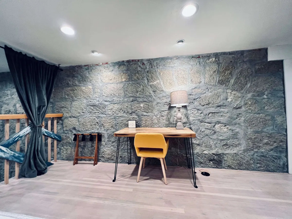 A cozy work nook with a rustic stone wall, wooden desk, yellow chair, and a softly glowing lamp. The scene invites creative reflection or quiet study, all set within a character-filled space.
